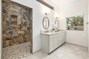 Modern bathroom with a glass-front shower clad in brown-gray slate tiles, pebble floor, and a white double vanity with oval mirrors.