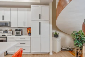 Bright white kitchen with tall pantry cabinets, stainless steel appliances, light wood floors, and a curved wooden spiral staircase on the right, with a few plants.
